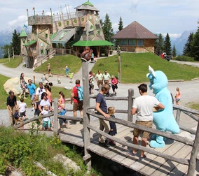 A group of visitors interact with a person in a blue dragon costume on a wooden bridge in Snow Space Salzburg, in front of an ornate play tower and green landscape