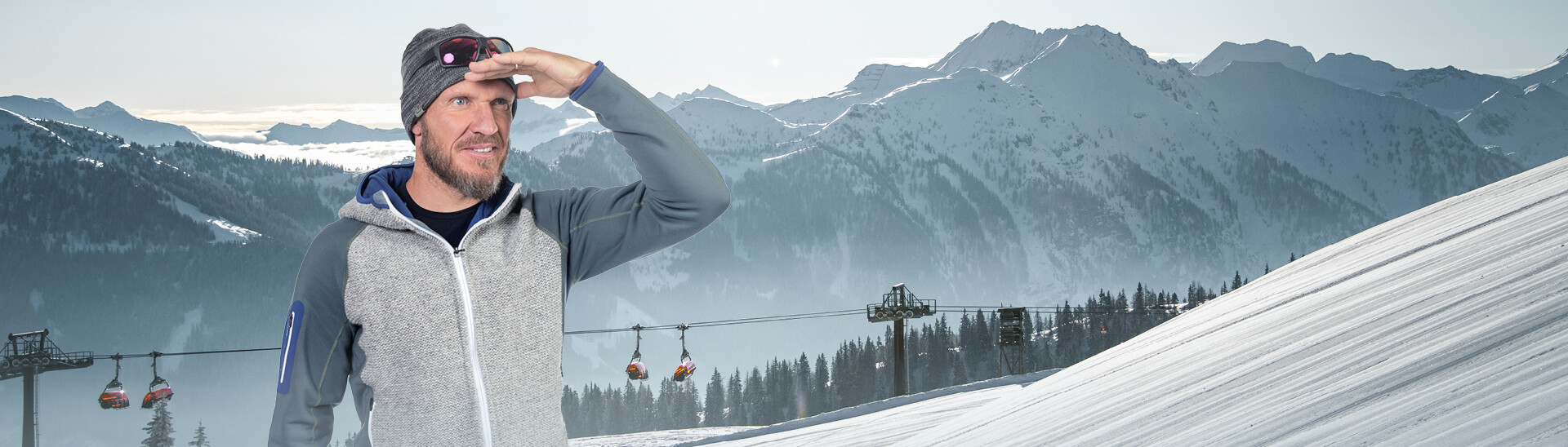 Hermann Maier in outdoor clothing stands in front of a wintry mountain landscape with cable car gondolas