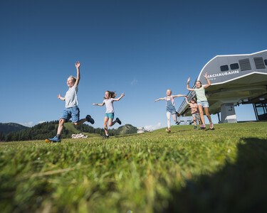 Four children jumping for joy across the meadow, behind them the Blackaubahn at Snow Space Salzburg in bright sunshine