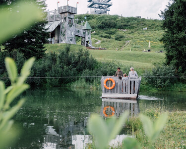Visitors on a wooden raft in an idyllic lake against the backdrop of a play tower in Snow Space Salzburg